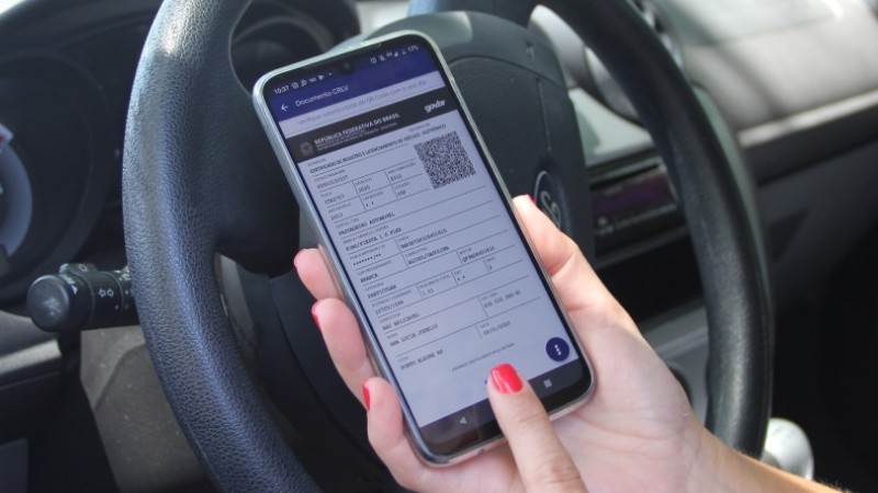 Imagem do documento na tela de um celular, segurado por uma pessoa diante de volante de veículo