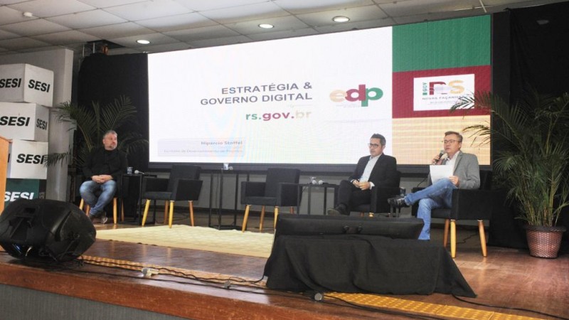 Três homens no palco diante de telão que exibe slide com o título "Estratégia & Governo Digital rs.gov.br" 