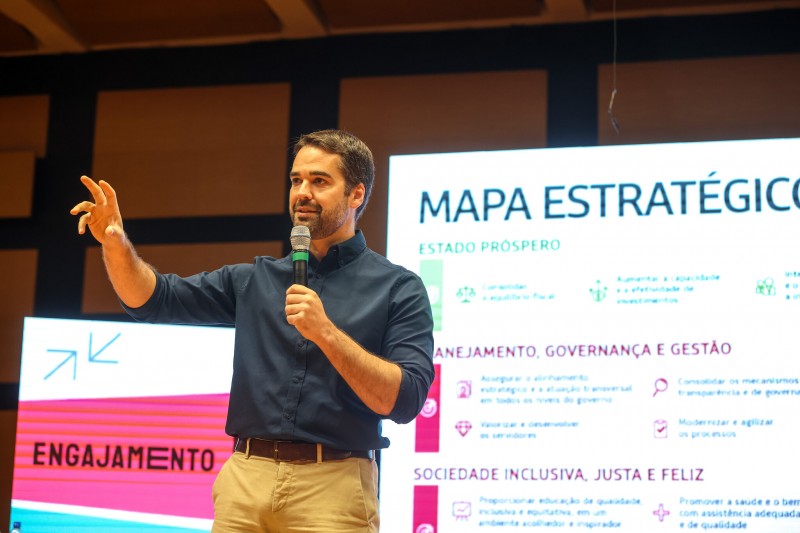 Governador Eduardo Leite discurso ao microfone em frente a tela que exibe o Mapa Estratégico do Governo