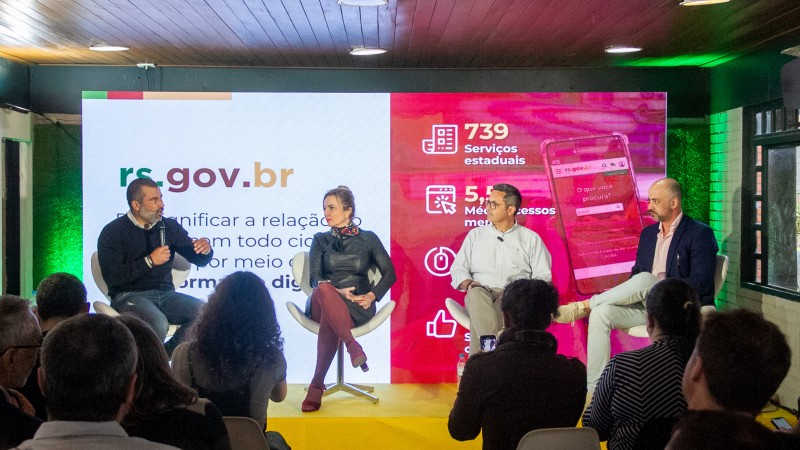 Silveira, Karen, Stoffel e Maraschin sentados em cadeiras, à frente de um painel com destaques do rs.gov.br, falam a plateia na Expointer