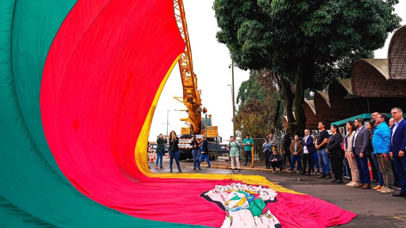 Varias pessoas perfiladas na frente da Ceasa/RS enquanto assistem Ao levantamento de uma bandeira gigantesca do Rio Grande do Sul que fica suspensa por um guincho.