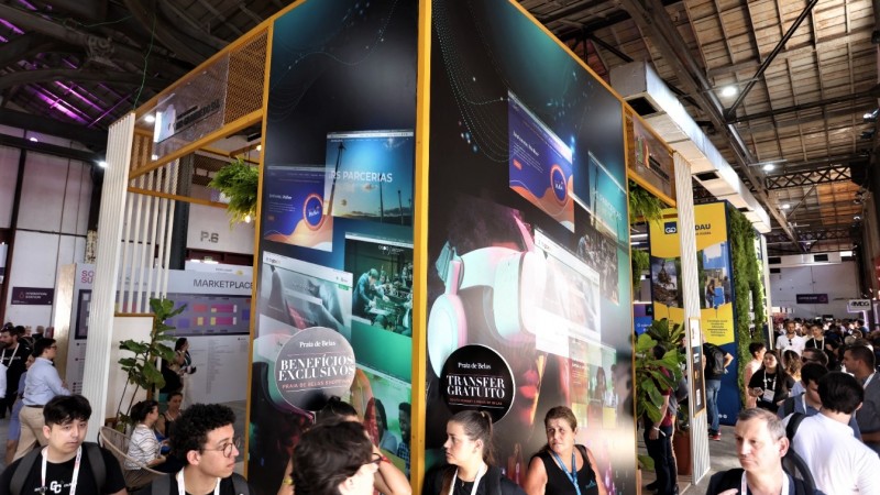 Imagem geral do estande global do governo do Estado no South Summit Brazil. Ele é visto de fora e está em destaque a estrutura toda adesivada com imagens que remetem à tecnologia e que forma uma quina de paredes delimitando o espaço. A estrutura é alta, em volta dela, circulam diversas pessoas.